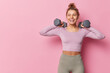 © WHstudio Leushin N - Athletic woman in sportswear raises dumbbells pumps biceps in gym being full of energy leads healthy lifestyle has happy expression isolated over pink background copy space for advertisement