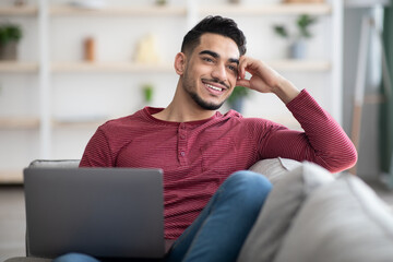 Wall Mural - Dreamy arab guy resting on couch at home, using laptop