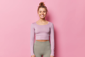 Happy delighted sporty woman in top and leggings going to do aerobics being in good physical shape smiles gladfully isolated over pink background. Fitness model in tracksuit ready for training