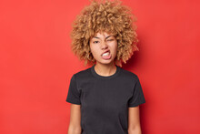Waisy Up Shot Of Irritated Curly Haired Young Woman Clenches Teeth Winks Eye Has Cheeky Expression Dressed In Basic Black T Shirt Isolated Over Red Background Being Dissatisfied With Something.