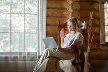 A Blonde Woman In Beige Home Clothes Sits In A Rocking Chair With A Laptop In Her Hands In A Cozy Room With Large Windows And Wooden Walls. Freelancing And Distance Learning.