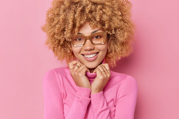 Wall Mural - Portrait of curly haired woman smiles gently keeps hands on collar of jumper expresses positive emotions hears good news wears spectacles isolated over pink background. Happy emotions concept