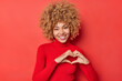 © WHstudio Leushin N - Feminine young happy woman with curly hair cheerful smile shows heart sign looks tenderly at camera expresses love to someone stands in turtleneck against vivid red background. Be my valentine