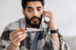 © Prostock-studio - Shocked ill arab man calling doctor while looking at thermometer, sitting on sofa wrapped in blanket, selective focus