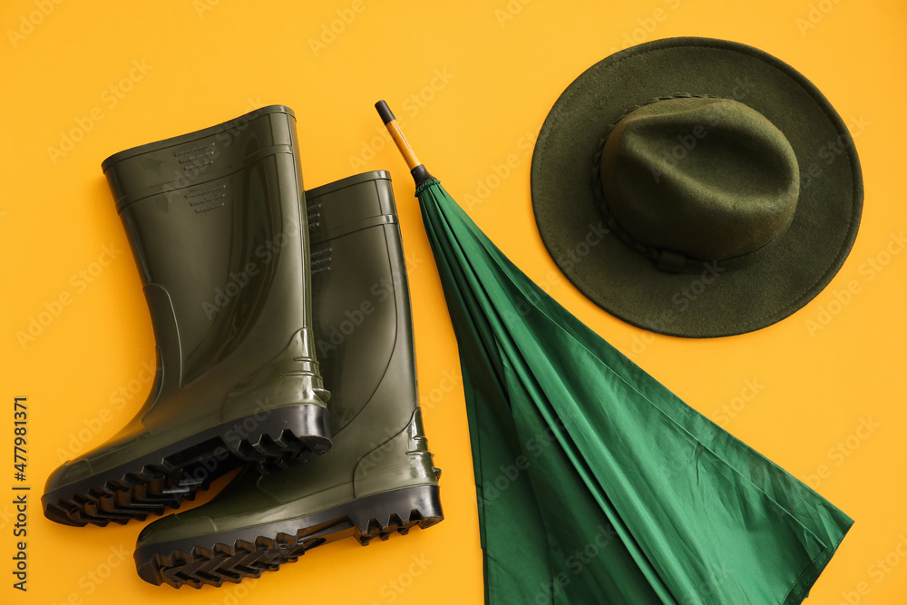 Umbrella, rubber boots and hat on color background