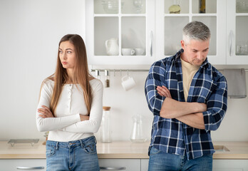 Offended mature married couple standing in kitchen with crossed arms after quarrel at home