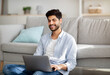 © Prostock-studio - Modern job lifestyle. Happy arab man working on laptop online, sitting on floor and leaning on sofa, copy space