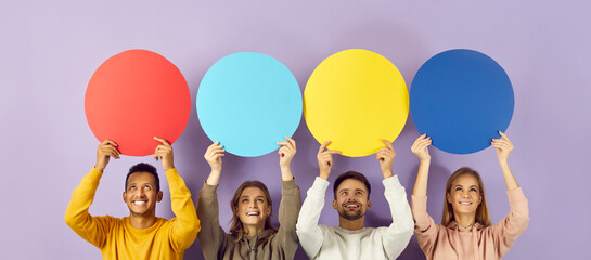 Studio portrait of happy cheerful positive young multiracial people sharing opinions or sending messages, holding round colourful red, blue and yellow blank mock up speech bubbles on purple background