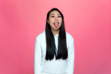 Crazy Playful Asian Young Girl Making Faces Showing Tongue, Having Fun, Teasing Fooling On Modern Pink Studio Background