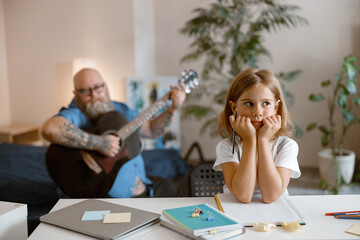 Wall Mural - Schoolgirl sits at table while daddy bothers her playing guitar on sofa in living room