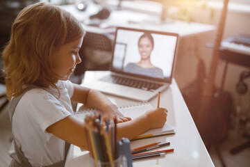 Wall Mural - Schoolgirl writes listening to teacher at videocall via modern laptop at table in light room