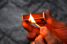 Detail Of Ignition Of Matches For A Matchbox. A Burning Match In One Hand And A Matchbox In The Other. The Process Of Lighting A Match. Hand With Burning Match On Gray Background