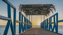 Dawn Over The Red Sea. A Wooden Pedestrian Walkway With Metal Railings Goes Over The Water. The Canopy Is Covered With Palm Leaves. The Rising Sun Turns The Sky Orange. Egypt