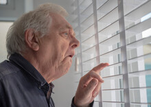 Senior Man With Funny Facial Expression Looking Out Of Window Blinds 