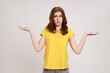 I don't know, who cares. Portrait of confused clueless attractive teenager girl in yellow t-shirt shrugging shoulders, making no idea gesture, whatever. Indoor studio shot isolated on gray background.