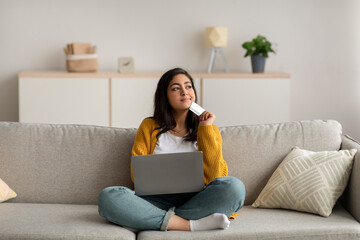 Wall Mural - Thoughtful middle eastern lady holding credit card and using laptop, dreaming about new clothes, purchasing via internet