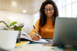 © Prostock-studio - Black woman sitting at desk, using pc writing in notebook