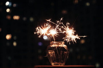 Wall Mural - Bengal lights in glass jar against background of  busy night city. Festive sparklers at night.