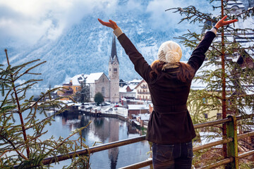 Wall Mural - A happy traveler woman in winter clothing enjoys the view to the village of Hallstatt in the Austrian Alps during winter time with snow