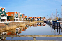 Panorama With Modern Brick Houses Along Water In A Family Friendly Suburban Neighborhood In Volendam In The Netherlands. Typical Dutch Houses And Windmill On Background.