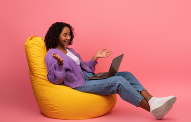 Sticker - Smiling young black woman making video call with friends, using laptop, sitting in bean bag chair on pink background