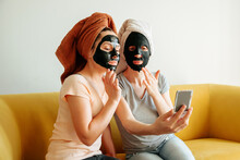 Two Girls With A Towel On Their Head And A Black Skin Care Mask. They Look In The Mirror And Touch Their Faces With Their Fingers.