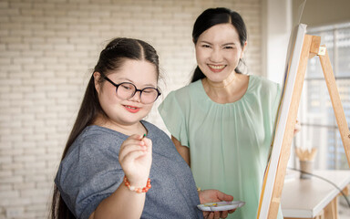 Wall Mural - Portrait shot of Asian happy lovely mother standing smiling with young chubby down syndrome autistic autism little daughter wearing eyeglasses holding paintbrush drawing painting on canvas easel.