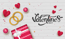 Happy Valentine's Day Font With Top View Of Golden Couple Rings, Gift Box, Paper Cut Hearts And Lit Tea Candles On White Wooden Texture Background.