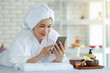 © Bangkok Click Studio - Asian happy relax female model in white clean bathrobe lay down on massage bed with compress ball coconut oil and plumeria flower in tray smiling using smartphone waiting for body massaging therapy