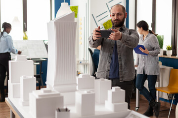 Poster - Architect working with smartphone to photograph white foam scale model maquette of skyscraper building complex. Engineer with mobile device next to desk in architectural modern office.