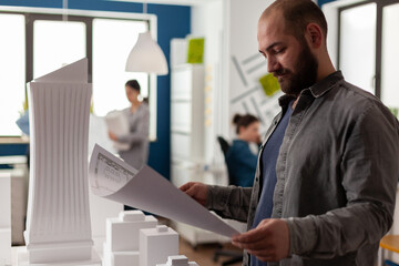Poster - Architect reading blueprint of residential project next to skyscraper maquette in modern architectural office. Engineer inspecting construction plans standing near desk with 3d foam model.