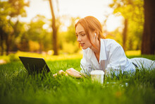 Young Redhead Student Girl Lyes On Grass Uses Laptop Pc And Headphones. Distance Education. 
