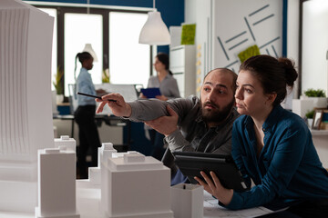 Poster - Architect pointing pen at skyscraper architectural model collaborating with colleague engineer holding tablet with blueprints in modern office. Architects looking inspired collaborating on maquette.