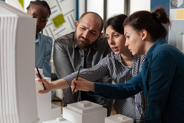 Poster - Two women professional architects explaining construction model design to team colleagues in front of white foam maquette. Engineers working together on residential project in architectural office.