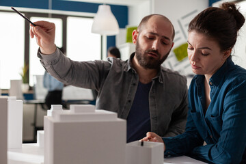 Poster - Architect looking at blueprints pointing pen at skyscraper architectural model collaborating with colleague engineer in modern office. Architects brainstorming ideeas collaborating on maquette.
