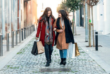 Two Beautiful Friends Talking And Having Fun While Walking On The City Street With Shopping Bag.