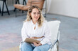 A blonde therapist with glasses is sitting on a white chair writing into a clipboard and smiling. Mental health of the patient. White shirt and blue trousers.