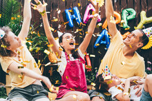 Joyful Family With Children Throws Air Balloons And Golden Confetti Sitting On Plaid At Birthday Celebration In Decorated Cottage Yard In Summer