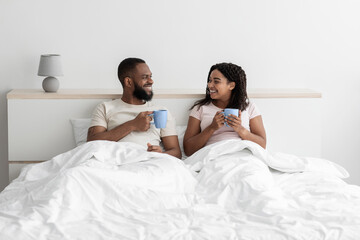 Wall Mural - Smiling happy millennial african american man and wife sit on bed in white bedroom interior with cups of coffee