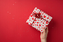 First Person Top View Photo Of St Valentine's Day Decor Female Hand Giving Giftbox In White Wrapping Paper With Heart Pattern Red Star Bow And Sequins On Isolated Red Background With Empty Space
