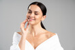 © NFstock - Portrait view of happy young caucasian woman touching her face and smiling with closed eyes, while standing in bathrobe isolated on grey copy space background. Skin and body care concept