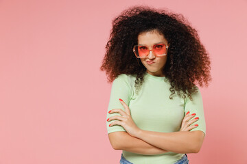 Wall Mural - Irritated dissatisfied frowning sad young curly latin woman 20s years old wear mint t-shirt sunglasses hold hands crossed looking camera isolated on plain pastel light pink background studio portrait