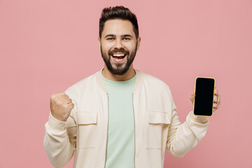 Wall Mural - Young smiling happy man 20s wearing trendy jacket shirt hold in hand use mobile cell phone browsing with blank screen workspace area do winner gesture isolated on plain pastel light pink background.