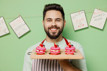 Close up young smiling happy male chef confectioner baker man 20s in striped apron hold three cake muffin macaroon on board isolated on plain pastel light green background studio Cooking food concept