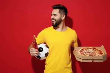 Wall Mural - Happy young man football fan in yellow t-shirt cheer up support favorite team hold soccer ball italian pizza in cardboard flatbox beer look aside isolated on plain dark red background studio portrait.