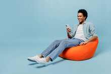 Full Size Body Length Smiling Young Black Curly Man 20s Years Old Wears White Shirt Sit In Bag Chair Hold In Hand Use Mobile Cell Phone Isolated On Plain Pastel Light Blue Background Studio Portrait.