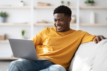 Joyful black guy sitting on couch with laptop