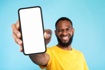 Wall Mural - Mobile app advertisement. Smiling black man showing smartphone with empty screen, winking on blue background, mockup