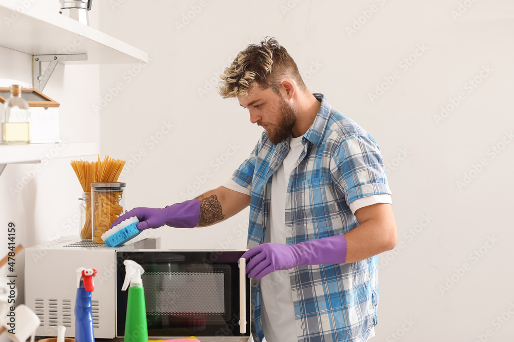 Handsome young man with sponge cleaning microwave in kitchen