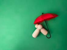Pair A Wooden Doll Fallen With A Red Umbrella On A Green Background With Copy Space Text. The Concept Of Insurance Coverage.